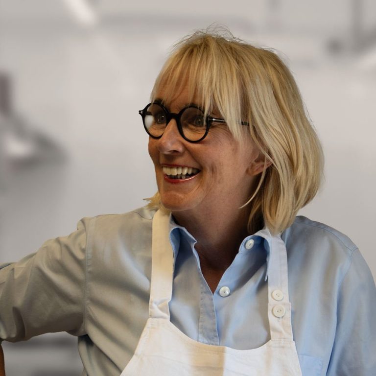 Martina Schwartz, eine lächelnde Frau mit blonden Haaren, Brille und Schürze in einer modernen Küche.